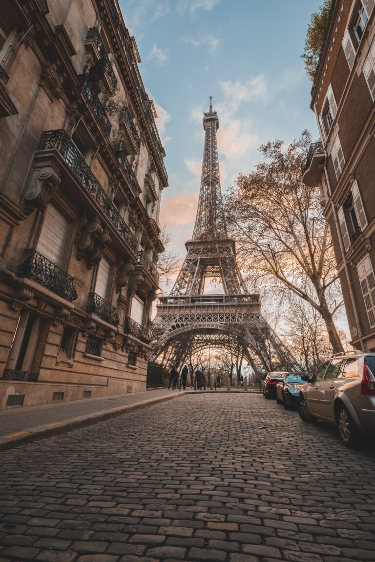 A sunset image of the Eiffel Tower, with a cloud-filled blue sky and cobbled street. 